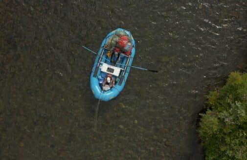 a downward aerial photo of an inflatable blue bloat floating on a river. the back of the boat is loaded with gear bags, and two people sit inside the boat, one rowing two oars