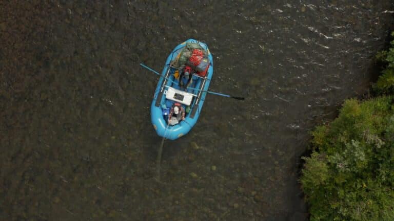 a downward aerial photo of an inflatable blue bloat floating on a river. the back of the boat is loaded with gear bags, and two people sit inside the boat, one rowing two oars