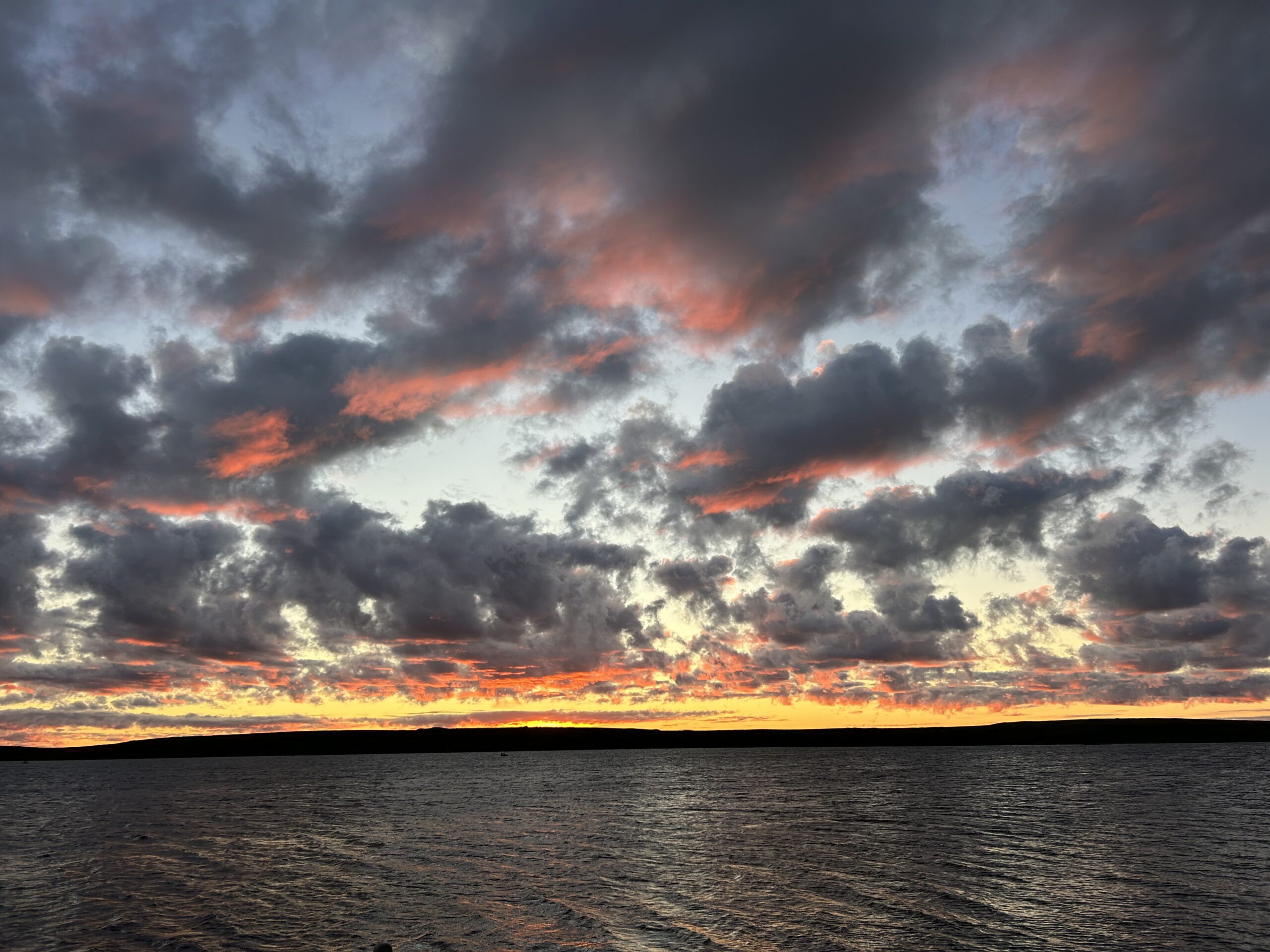 a cloudy sunset over water, dark grey clouds highlighted with a deep bright pink. a stripe of orangey yellow sky sits at the horizon