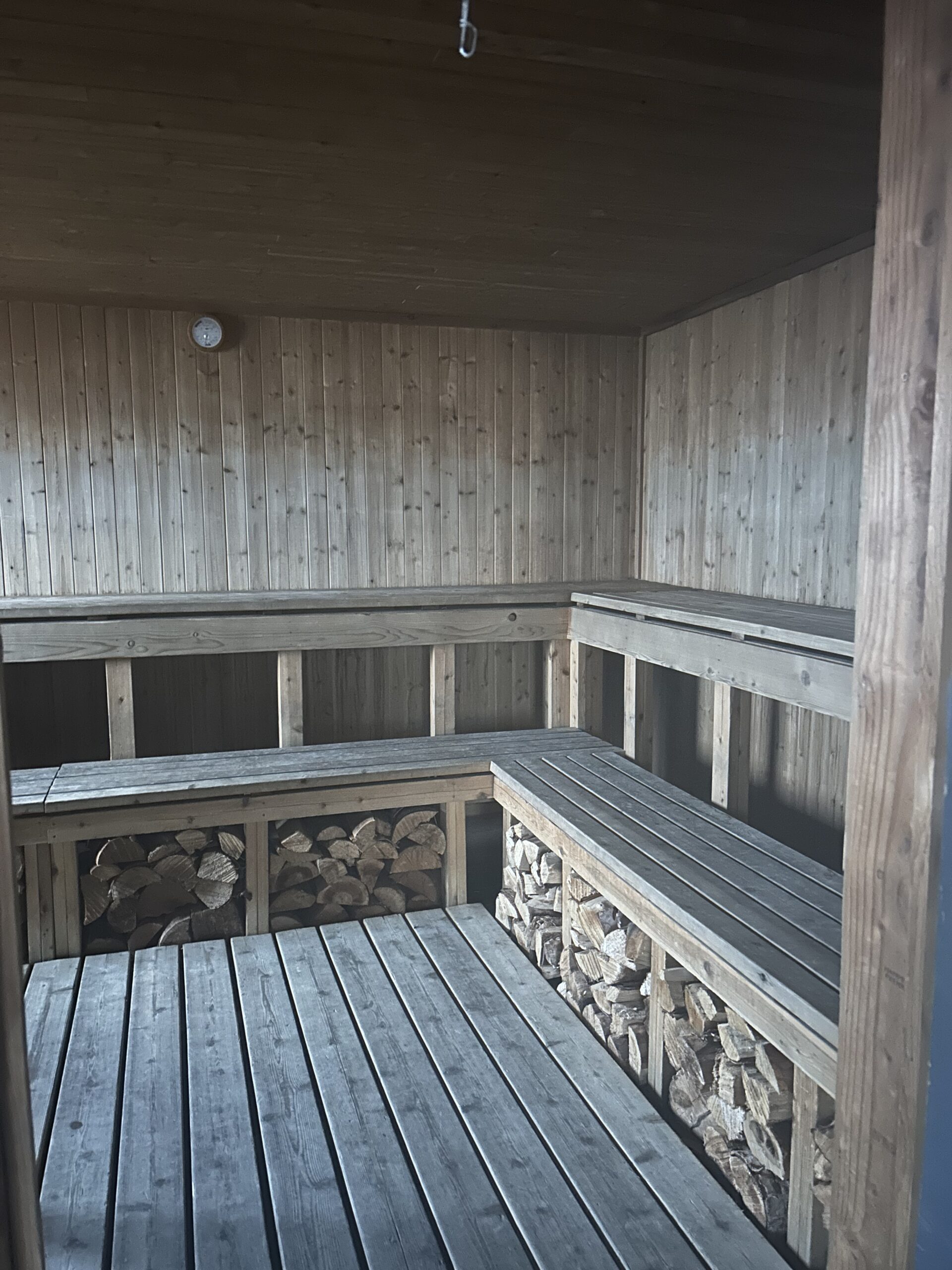 a wooden sauna with two levels of benches. Chopped wood is packed underneath the lower bench, all the way around the room