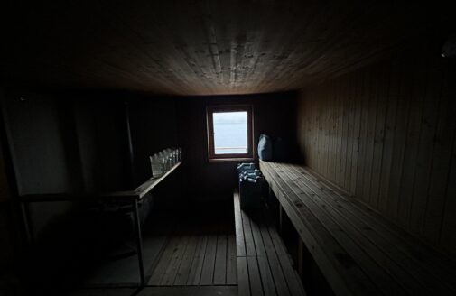 a dim photo of a wooden sauna. A window at the center of the frame provides a glimpse at the bright, overcast outdoors, and also sheds a limited amount of light into the room