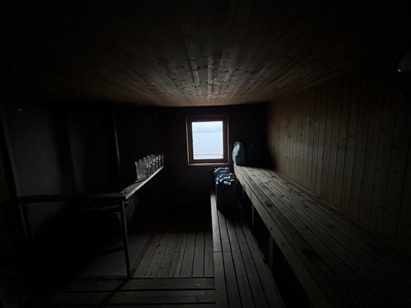 a dim photo of a wooden sauna. A window at the center of the frame provides a glimpse at the bright, overcast outdoors, and also sheds a limited amount of light into the room
