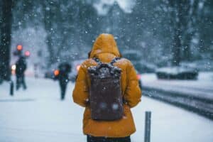 a person in a yellow winter coat and backpack walks away from the camera, down a snowy sidewalk, while snow falls