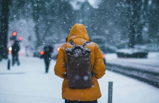 a person in a yellow winter coat and backpack walks away from the camera, down a snowy sidewalk, while snow falls