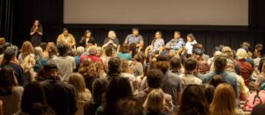 a seated panel of 7 speakers next to a moderator, on stage in front of a screen that says "MOUNTAINFILM." the audience is seated in an auditorium, applauding