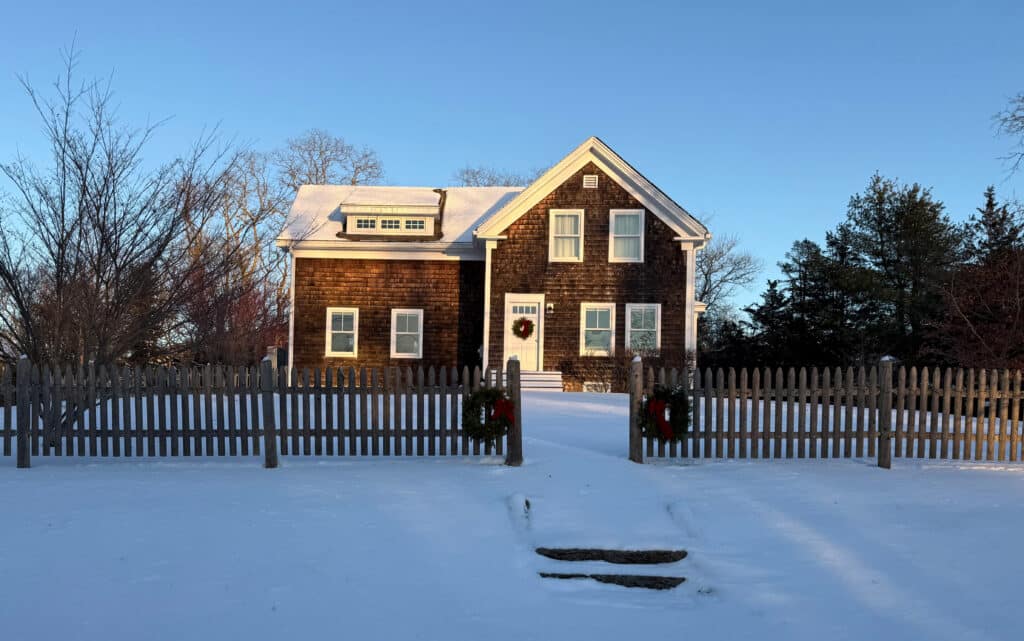 a new-england style house with brown wood shingle siding. the lawn and roof are covered in snow, and there is a picket fence