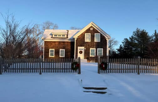 a new-england style house with brown wood shingle siding. the lawn and roof are covered in snow, and there is a picket fence