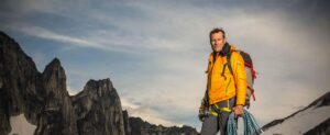 Conrad Anker stands on a rocky summit holding climbing ropes and wearing a backpack